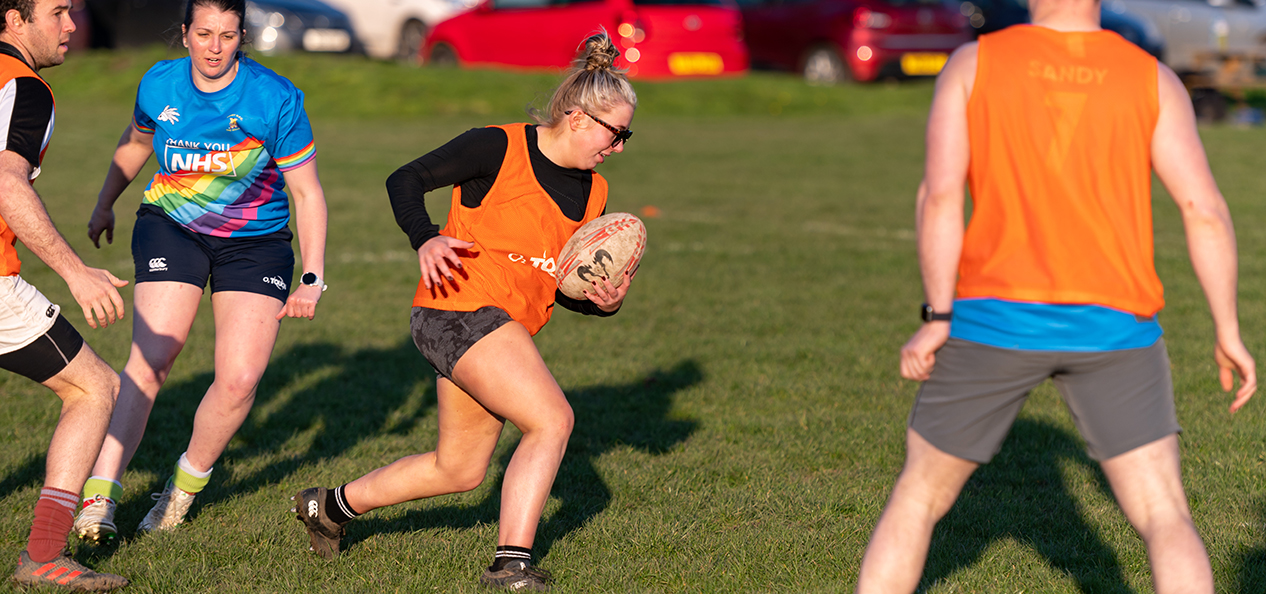 A woman runs as players from both teams look at her during an outdoors rugby game.