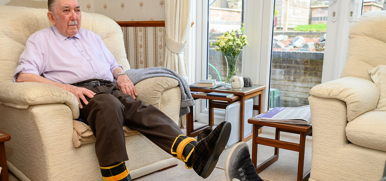 An older man performs seated exercises from his arm chair, wearing ankle weights.