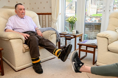 An older man performs seated exercises from his arm chair, wearing ankle weights.