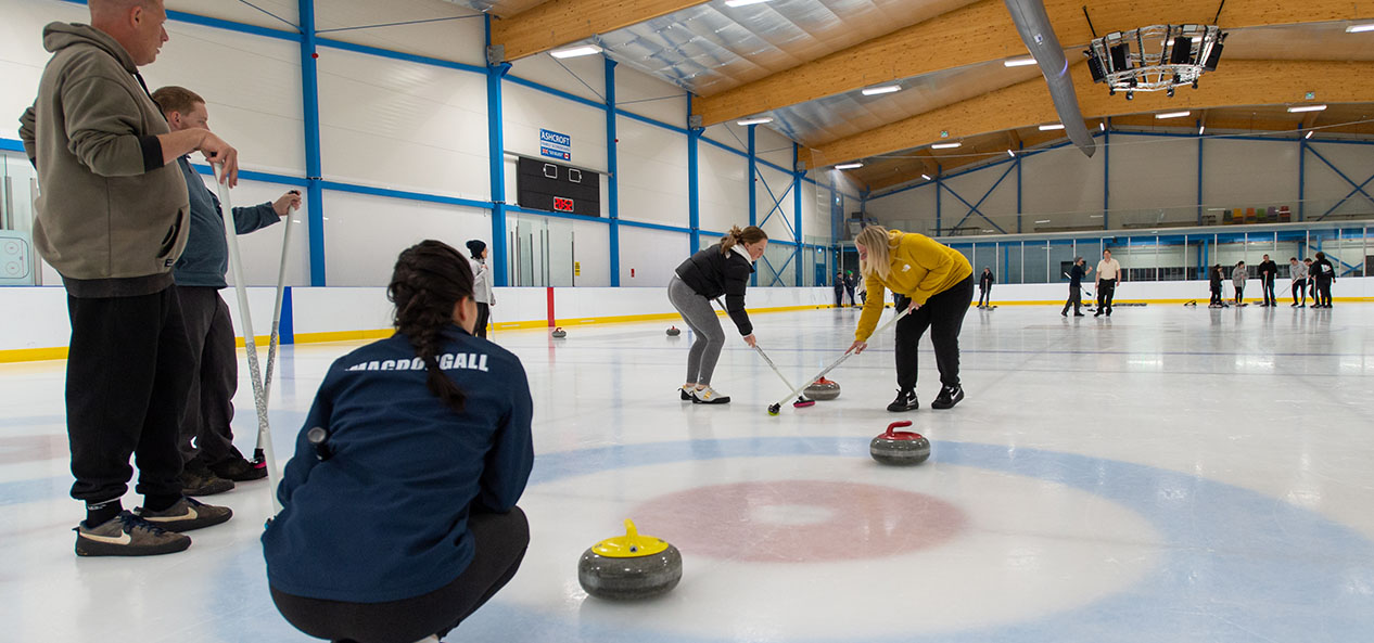 Two women sweep a curling stone as another crouches in the 'house' and two men watch on, also standing on the ice