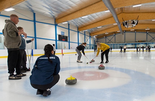 Two women sweep a curling stone as another crouches in the 'house' and two men watch on, also standing on the ice