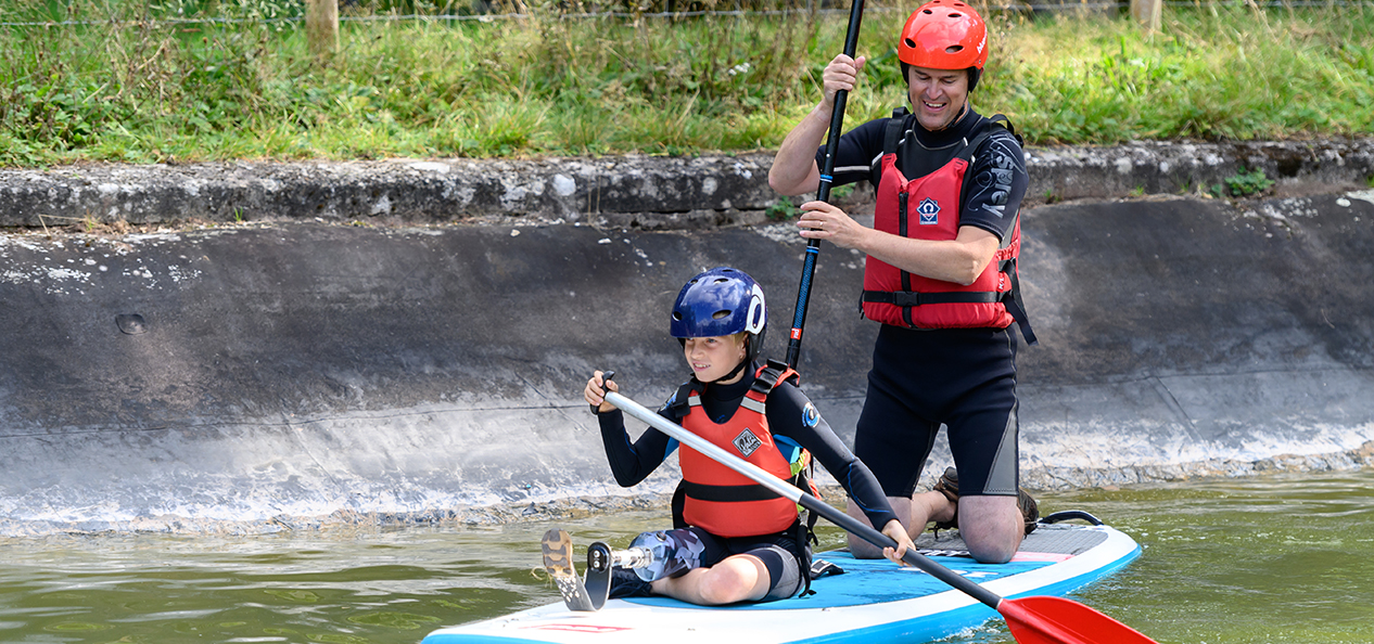 A boy with a prosthetic leg stands on a paddle board, on water. while an instructor kneels behind him