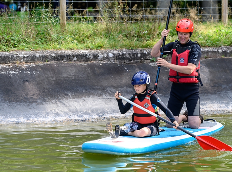 A boy with a prosthetic leg stands on a paddle board, on water. while an instructor kneels behind him