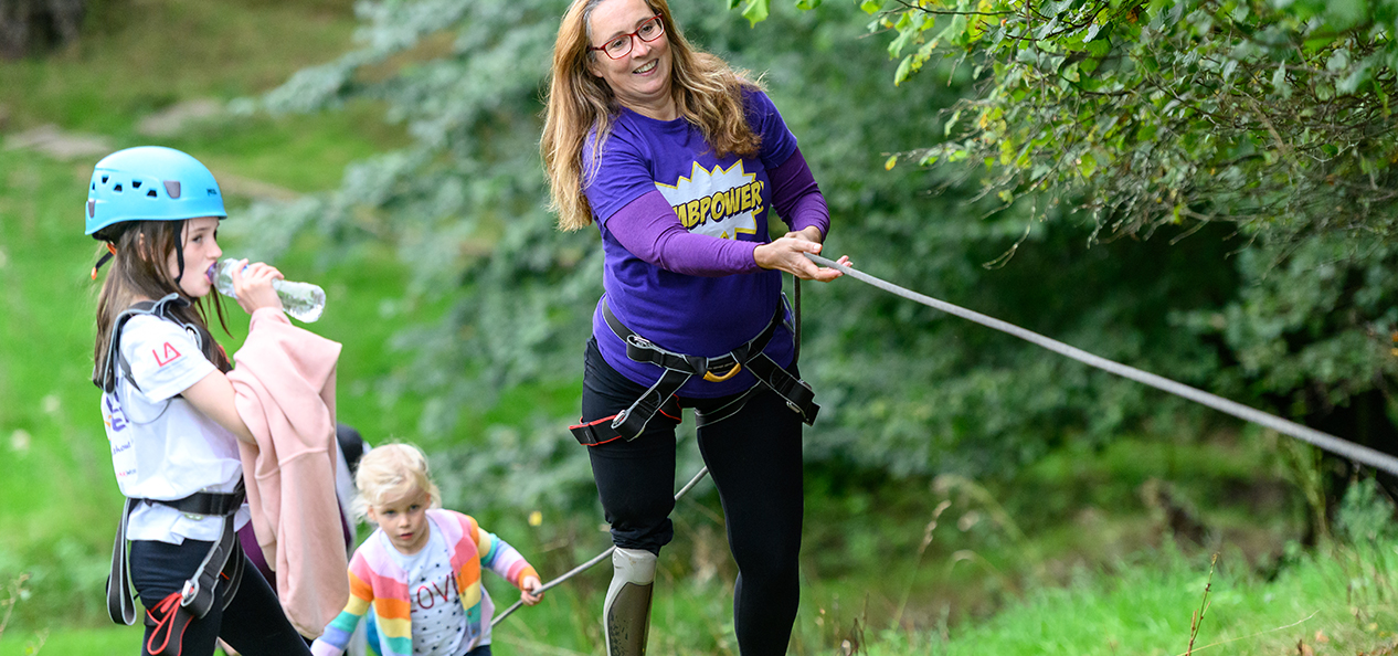 A woman with a prosthetic leg uses a rope to climb up a slope and two small girls watch on.