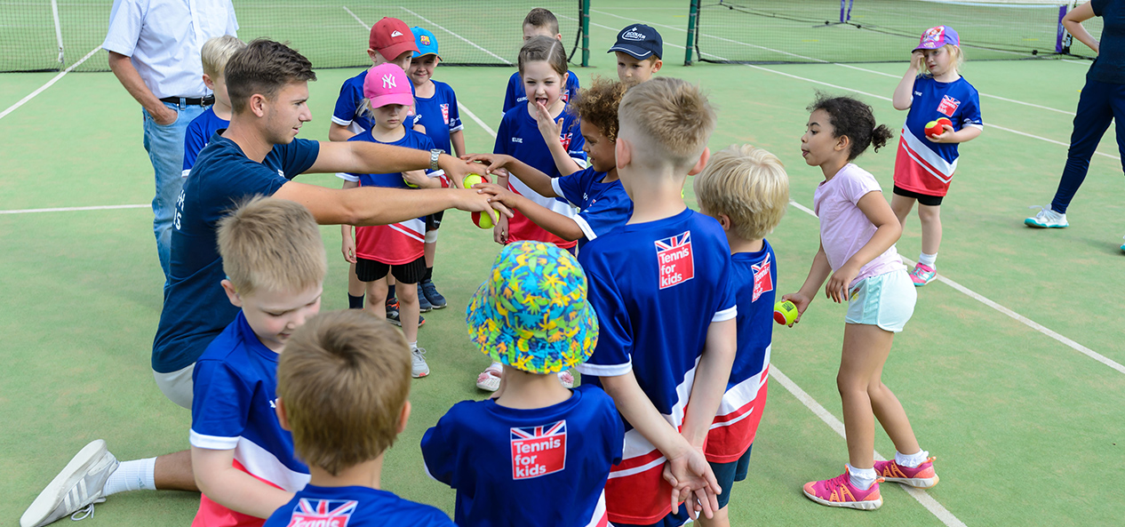 A tennis coach plays a game with a group of young children gathered around him