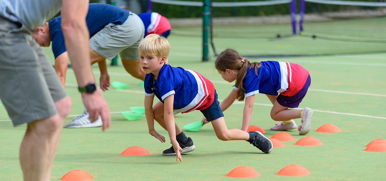 A boy and a girl train on an open-air tennis field under the supervision of their coaches.