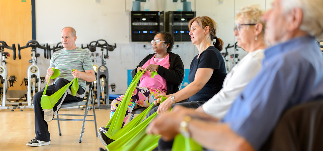 A group of senior men and women stretch with the help of some elastic bands on an indoor gym.