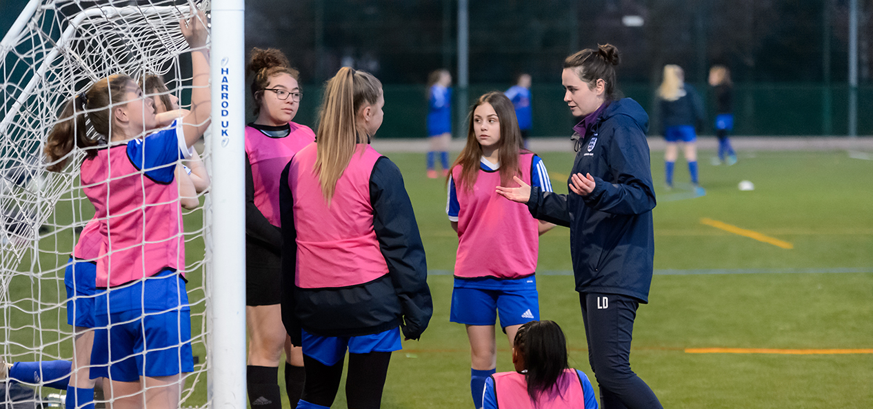 A female coach speaks to a teenage girls' football team at the side of an artificial football pitch