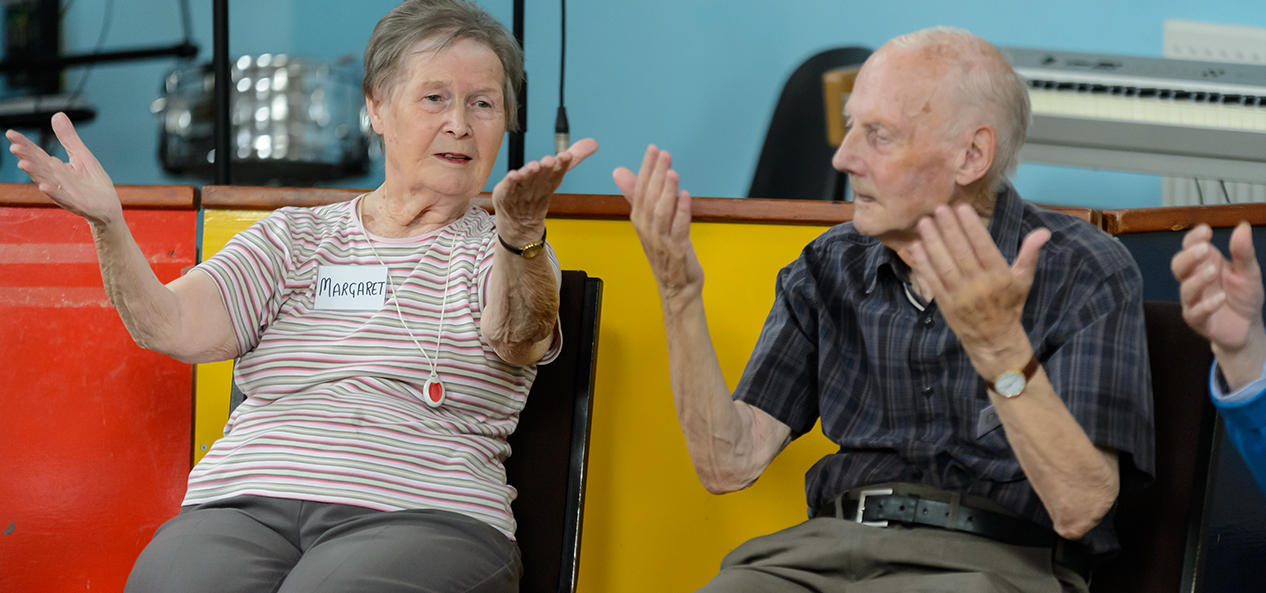 An old lady and an old gentleman perform stretching exercises while sitting on a chair indoors