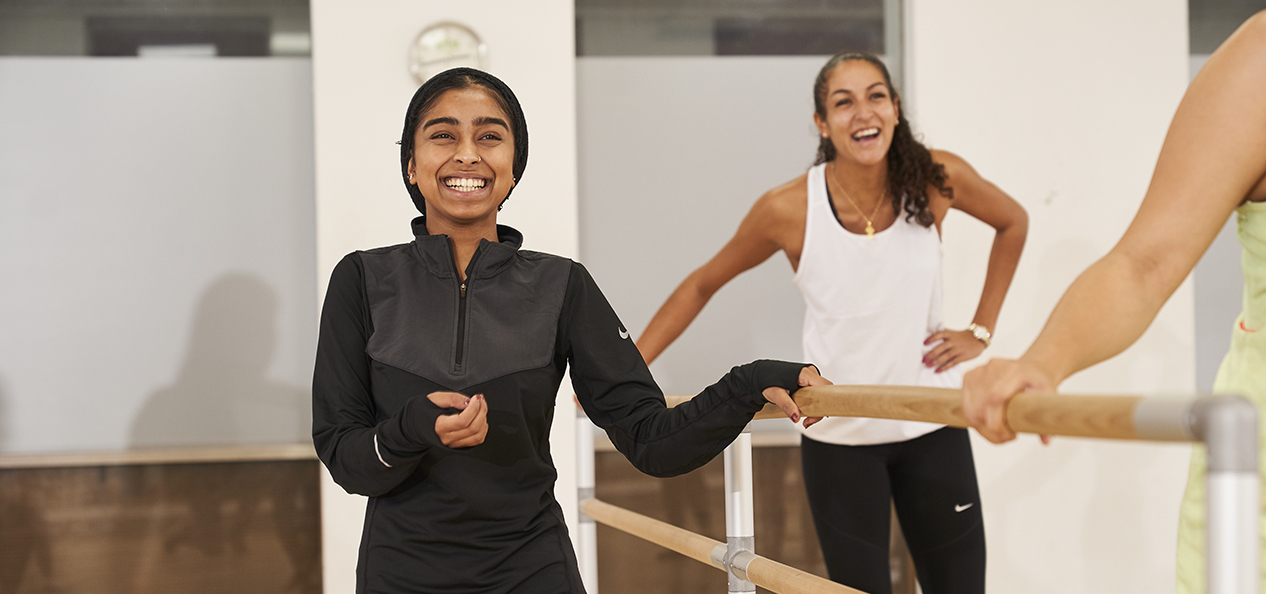 A smiling woman stands with one hand on a ballet barre, while another in the background also smiles and laughs