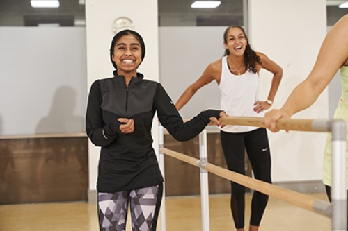 A smiling woman stands with one hand on a ballet barre, while another in the background also smiles and laughs