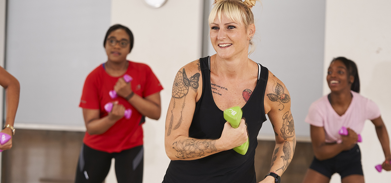 A group of women smile and exercise in a gym while holding small weights. 