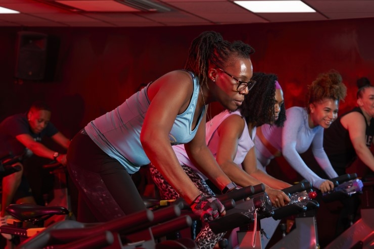 A row of women stand up while exercising on spin bikes in a studio.