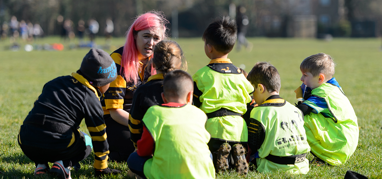 A female rugby coach crouches down with a group of children.