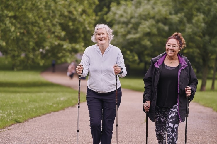 Two smiling older women walking in a park with walking sticks.