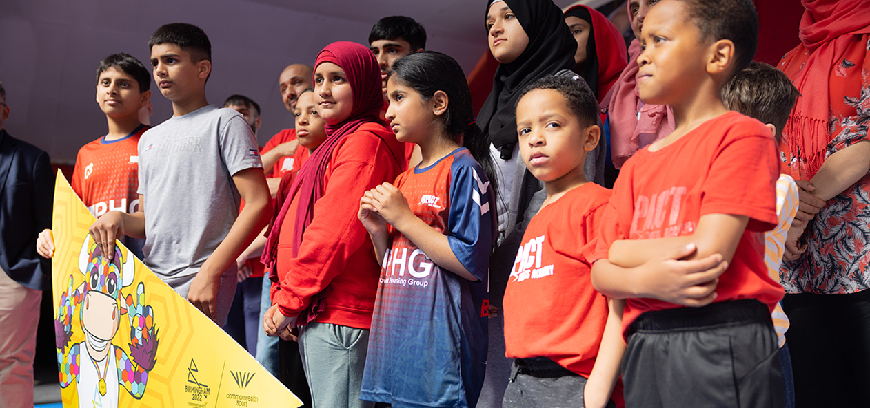 A group of children and women pose with a sign to celebrate the anniversary from the Birmingham Commonwealth Games 2022. 