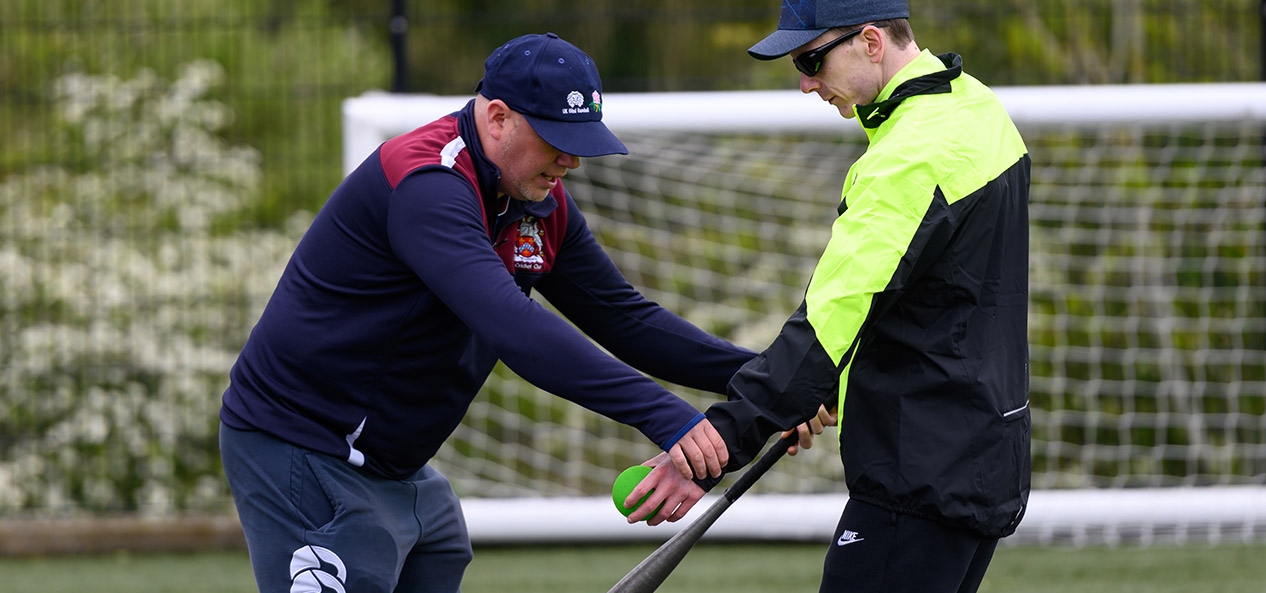 An instructor assists an visually impaired man to hit a tennis ball with a softball bat.