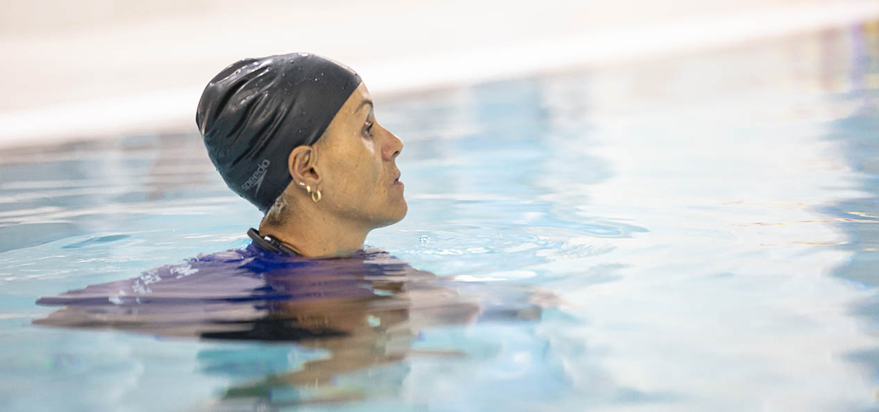 A black woman wearing a swimming cap looks ahead while in the water of an indoors swimming pool. 