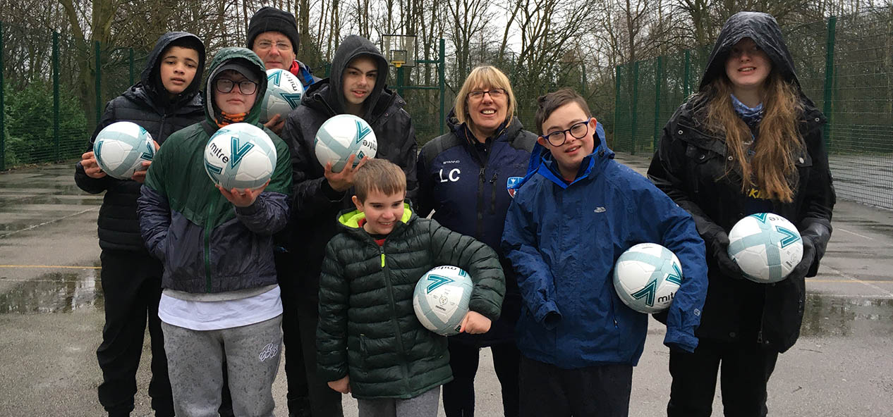 A group of disabled young people, with a coach, stand in a playground holding footballs as they pose for the camera.