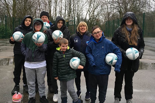 A group of disabled young people, with a coach, stand in a playground holding footballs as they pose for the camera.