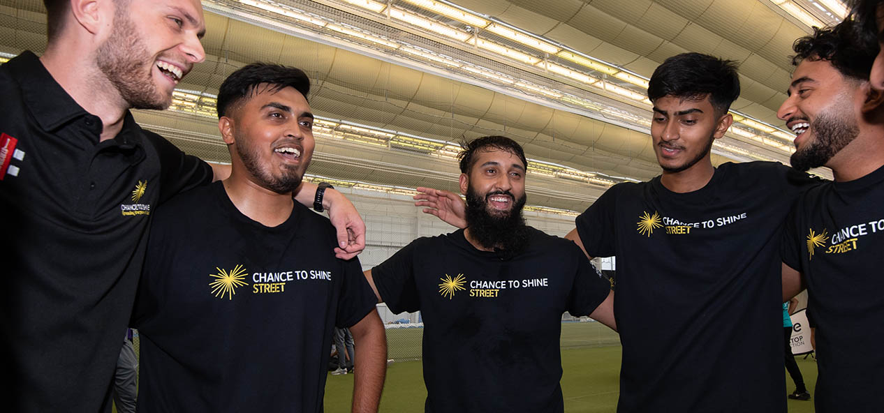 A group of men wearing Chance to Shine Street t-shirts stand on a circle huging each other and smiling. 