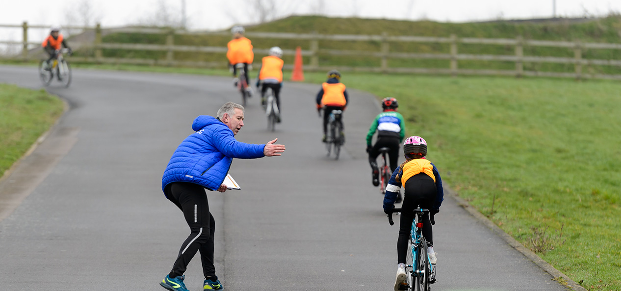 A volunteer instructing a group of children as they ride past.