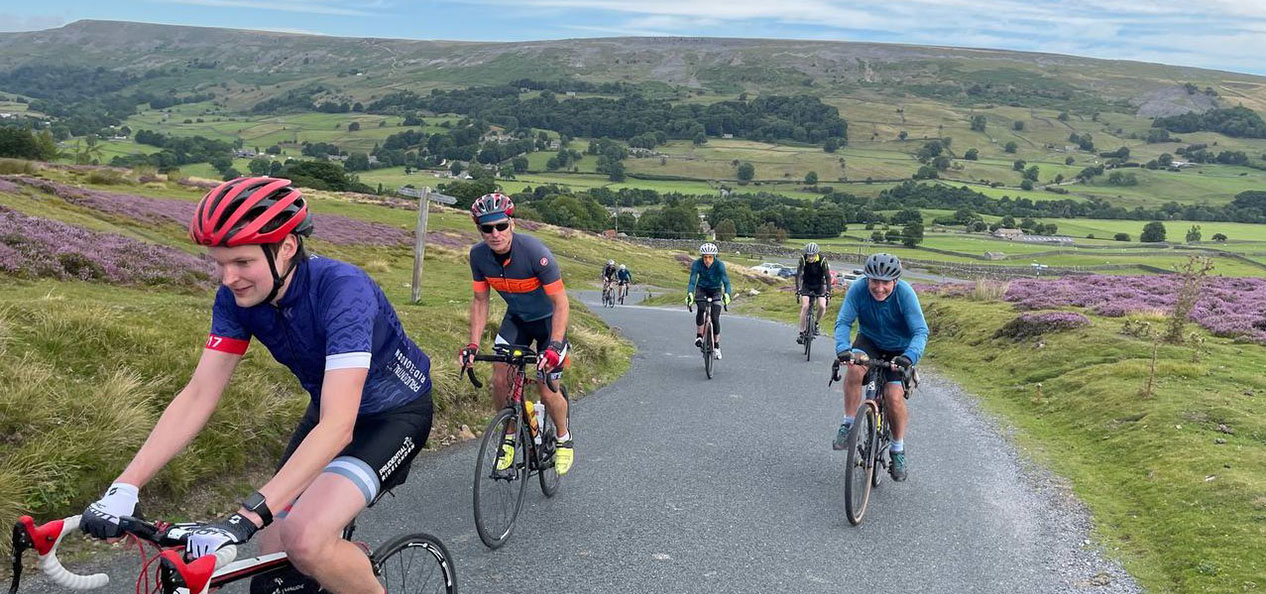 A group of men on bikes and wearing helmets, gloves and other cycling gear cycle up a hill.