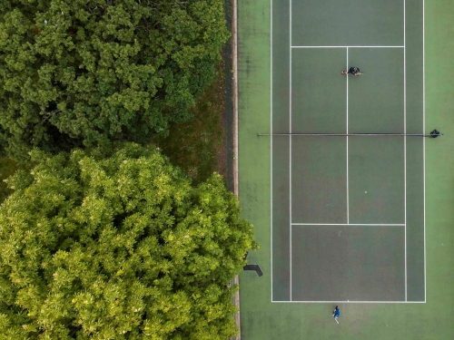 An overhead shot of a green tennis court in a park, next to green trees.