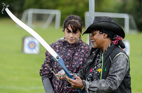 A female archery coach instructs another woman at an archery lesson outdoors.