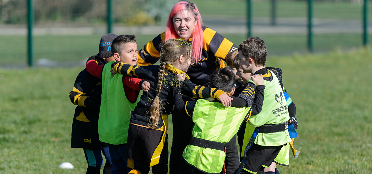 A rugby coach speak to a group of children who have formed a huddle around her.