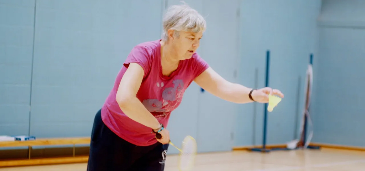 An older woman prepares to serve while playing badminton in a sports hall