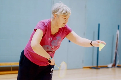 An older woman prepares to serve while playing badminton in a sports hall