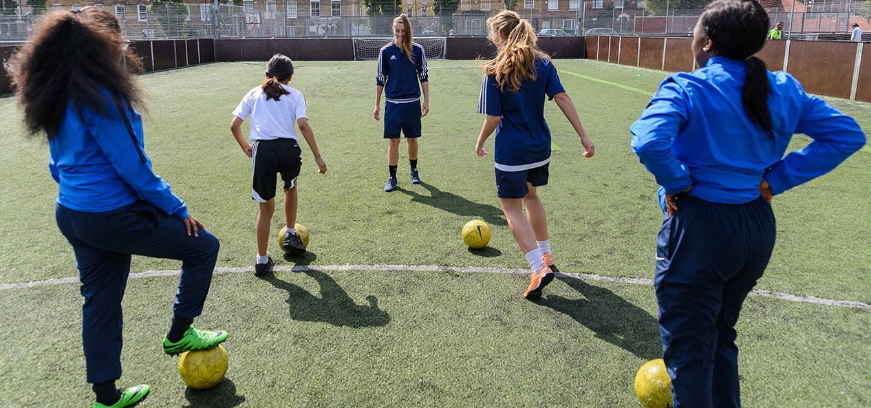 A coach in front of a group of women standing in front of footballs on a pitch.